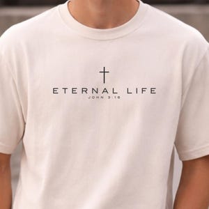 Puede incluir: Una camiseta de manga corta color crema con un gráfico negro en el pecho. El diseño incluye un símbolo de cruz simple sobre el texto ETERNAL LIFE y el texto más pequeño JOHN 3:16 centrado debajo de la frase principal en una fuente clara.