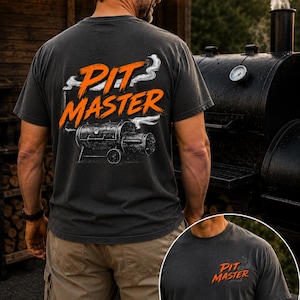 May include: Dark gray t-shirt with the words "Pit Master" in orange and white, along with a graphic of a smoker. The shirt is worn by a person standing near a large black smoker.