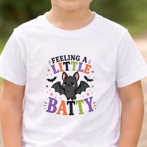 May include: A white short-sleeved t-shirt featuring a graphic of a cartoon bat with the text "FEELING A LITTLE BATTY" in orange, purple, and green. The design includes small stars and two smaller black bats surrounding the central character.