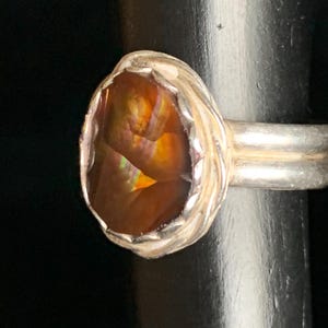 May include: A silver ring featuring an oval-cut gemstone with fiery orange, yellow, and red hues. The gemstone is set in a textured bezel, and the ring has a double-band design. The ring is displayed against a dark background.