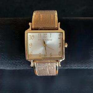 May include: A gold-toned rectangular wristwatch with a textured, light-colored face. The watch features gold hands and hour markers, with the brand name "Betsey Johnson" printed on the face. The watch has a gold band with a textured pattern.