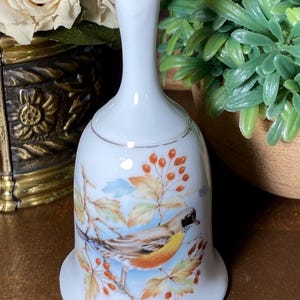 May include: A white porcelain bell featuring a hand-painted illustration of a bird with a yellow and orange breast perched on a branch with orange berries and light-colored leaves. The bell has a gold-toned rim at the base and a gold-toned handle top.