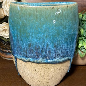 May include: A handmade ceramic vase with a unique design. The top half is glazed in shades of turquoise and blue, with a dripped effect. The bottom half is a natural, speckled tan color. The vase has a smooth, rounded shape.