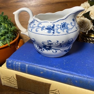 May include: White ceramic creamer with blue floral patterns. The creamer has a curved handle and a spout for pouring. It is sitting on a blue book. The creamer is a vintage piece, perfect for serving cream or milk.