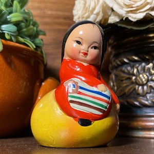 May include: A vintage ceramic figurine of a seated doll with black hair, wearing a red outfit and a colorful apron. The doll is perched on a yellow base. The figurine has a painted face with rosy cheeks and red lips.