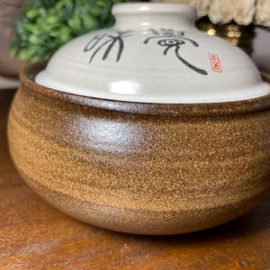May include: A brown ceramic bowl with a lid. The bowl has a textured surface and a light-colored lid with black calligraphy and a red stamp. The bowl is likely used for serving food.
