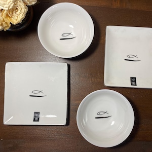 May include: A set of four white ceramic dishes with a minimalist fish design. The set includes two square plates, and two bowls. Each piece features a black fish illustration and a small black square symbol.