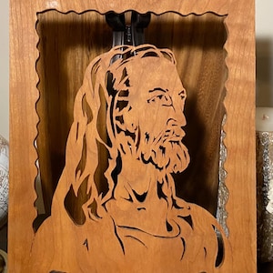 May include: A wooden relief carving of a religious figure, framed in a rectangular wooden border. The figure has long hair and a beard, with the carving showing intricate details. The wood has a warm, natural tone.