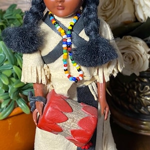 Può includere: Una bambola vintage vestita con abiti nativi americani. La bambola ha la pelle scura, trecce nere e una fascia per la testa con perline. Indossa un vestito color crema e porta una pochette rossa e beige. Una collana di perline colorate aggiunge un tocco di colore.