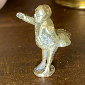 May include: A small, metallic, gold-toned figurine depicting a standing figure with a rounded torso, wearing a coat with tails. The figure is posed with one arm extended forward and the other arm bent at the side, all mounted on a circular base.