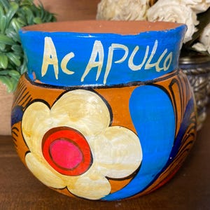 May include: A round terracotta pot featuring a wide blue band at the top with the word Acapulco painted in white. The body of the pot is orange and decorated with a large cream-colored flower with a bright red center and blue leaf accents.