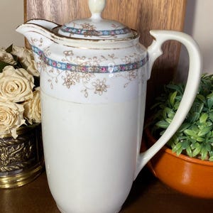 May include: A vintage white porcelain coffee pot with a lid. The pot features a decorative band of blue, pink, and gold floral patterns around the top. It has a curved handle and a spout for pouring. The pot is a classic piece of kitchenware.