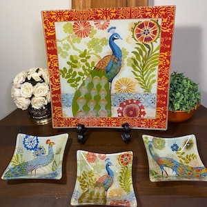 May include: A decorative peacock-themed glass serving set. The set includes a large square platter and three smaller square dishes. The peacock design features blue, green, and orange colors with floral accents. The platter has a red border.