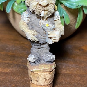 May include: A hand-carved wooden bottle stopper featuring a bearded figure wearing a dark cap and vest. The figure holds a small goblet with a yellow accent in one hand and has a white object in its mouth. The base is a cylindrical cork.