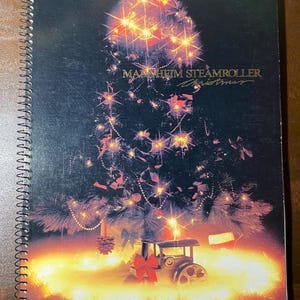 May include: A spiral-bound music book titled Mannheim Steamroller Christmas, PS-1984, on American Gramophone Records. The cover features a dark background with a lit Christmas tree, a toy steamroller with a red bow, and glowing warm lights.
