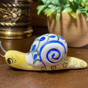 May include: A whimsical ceramic snail figurine. The snail's shell is white with blue and green painted details. The body is yellow with black flower accents and a smiling face. The snail's antennae are silver.