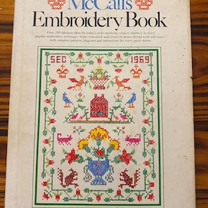 May include: A softcover book titled McCall's Embroidery Book with a white cover featuring a large, colorful cross-stitch sampler design. The sampler includes motifs of a house, birds, flowers, and animals in red, blue, yellow, and green.