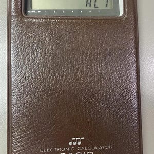 May include: A brown, rectangular Casio electronic calculator with a textured, leather-like finish. The device features a digital LCD screen displaying the text AL 1, along with labels for TIME SIGNAL, DATE MEMORY, and ALARM, plus a musical note icon.