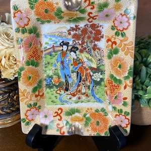 May include: Square ceramic ashtray with a beige background and a painted scene of two figures in traditional Asian attire. The border features floral designs in orange, pink, and green. The ashtray is supported by a black stand.
