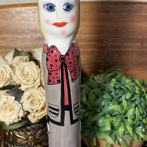 May include: A tall, ceramic figurine of a woman with blonde hair, blue eyes, and red lips. She wears a gray outfit with a red polka-dot bow tie. The figurine is on a black base.
