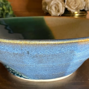 May include: A handmade ceramic bowl with a wide, shallow design. The bowl features a gradient of colors, including blue, green, and tan, with a golden rim. The interior is a deep green, transitioning to blue and tan. This bowl is perfect for serving.