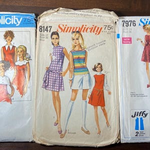 May include: Three vintage Simplicity sewing pattern envelopes. The envelopes feature illustrations of women in various dresses and outfits. The patterns are labeled with numbers and the Simplicity logo. The envelopes are in various states of wear.