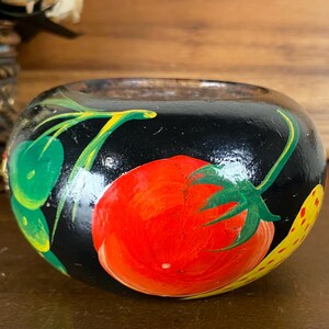 May include: A small, round, black wooden bowl featuring hand-painted illustrations of a bright red tomato with green leaves and stems, alongside green foliage and a yellow patterned element on the side of the smooth, glossy exterior surface.