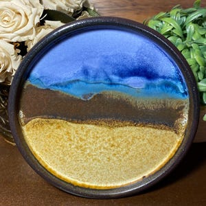 May include: A round ceramic plate with a coastal design. The plate features a blue sky, brown land, and a yellow beach. The rim is dark brown. The plate is a decorative piece for the home.