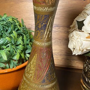 May include: A vintage brass vase with an etched design and colorful accents of red, blue, and yellow. The vase has a slender, hourglass shape and a wide base. Decorative home decor item.