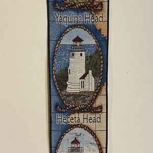 May include: A decorative wall hanging featuring three lighthouse images: Yaquina Head, Heceta Head, and Cape Arago. Each lighthouse is framed in a rope-like border against a blue and tan background. The bottom has a compass rose design.
