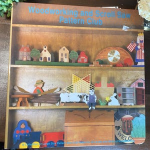 May include: A book titled Woodworking and Scroll Saw Pattern Club featuring a cover image of wooden shelves displaying various handcrafted items including miniature houses, a clock, a star, a boat, sheep, a basket, and a toy train.