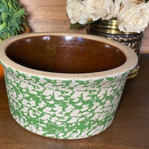 May include: An oval-shaped, antique stoneware bowl with a speckled green and cream exterior. The interior is a rich brown color. The rim is a light cream color. The bowl is likely used for serving or storage.