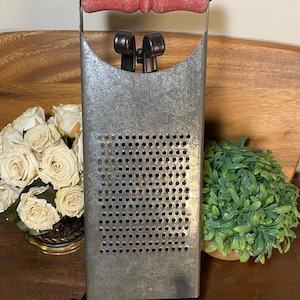 May include: A rectangular metal kitchen grater featuring a weathered silver finish and a cylindrical wooden handle painted in a distressed red. The grater has a flat surface with a grid of small circular perforations for shredding food items.