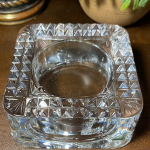 May include: A clear, square glass ashtray with a central circular depression. The ashtray features a geometric, textured design around the rim and sides, creating a sparkling effect. The ashtray is designed to hold cigarettes or cigars.