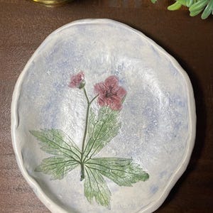 May include: A round, hand-formed ceramic dish with an irregular, wavy rim. The surface features a mottled, light blue and white wash finish. A detailed botanical illustration of a pink flower with green leaves is centered on the dish.