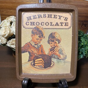 May include: A brown rectangular tray with a vintage Hershey's Chocolate advertisement. The image features two children and a chocolate cake. The tray is displayed on a black metal stand.