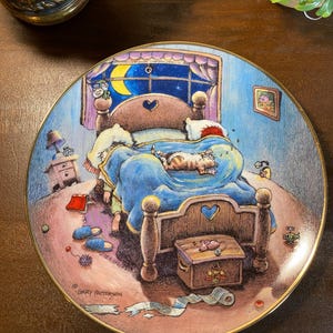 May include: A round decorative plate featuring a colorful illustration by Gary Patterson of a bedroom scene at night. A cat sleeps on a blue blanket atop a wooden bed, with a crescent moon visible through the window and scattered items on the floor.