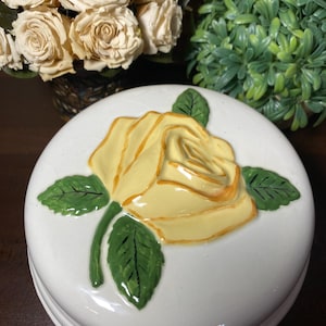 May include: A white ceramic trinket box with a raised yellow rose design and green leaves. The lid is round and the box is set against a background of flowers and greenery.