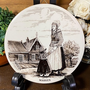May include: A round, off-white decorative plate featuring a sepia-toned illustration of a woman and child in traditional Dutch clothing, standing in front of a house. The word "MARKEN" is printed at the bottom. The plate is displayed on a black stand.