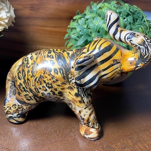 May include: A ceramic elephant figurine with a unique animal print design. The body features a leopard print pattern, while the trunk and ears have a zebra stripe design. The figurine is a decorative item.