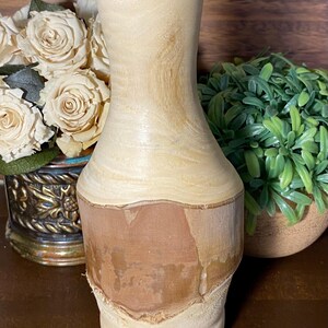 May include: A handcrafted wooden vase with a light beige upper section and a reddish-brown lower section. The vase has a smooth, curved design and a natural, unfinished look. The vase is a decorative item.