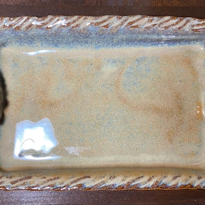 May include: Rectangular ceramic serving tray with a textured surface and a light beige and blue glaze. The edges have a decorative rope-like design and scroll-shaped handles.