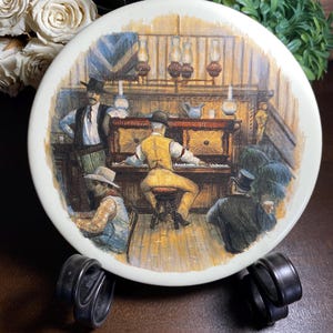 May include: A round, off-white decorative plate featuring a detailed illustration of a saloon scene. The image depicts several men, one playing a piano, with others observing. The plate is displayed on a black metal stand, with white roses in the background.