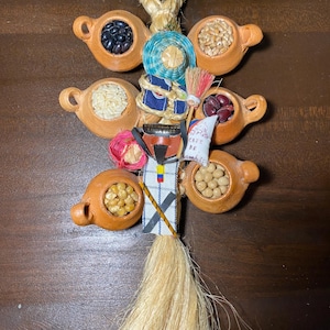 May include: A decorative hanging ornament featuring small terracotta pots filled with various grains and beans. The ornament includes a woven blue and white centerpiece, a small brush, and a tassel. The text on the ornament is in Spanish.