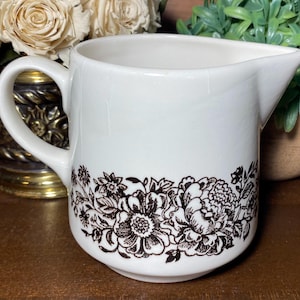 May include: White ceramic creamer with a curved handle and spout. The creamer has a floral design in brown along the bottom. A classic piece for serving cream or milk.