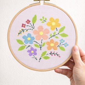 May include: A cross-stitch floral design in a wooden embroidery hoop. The design features colorful flowers in shades of yellow, orange, blue, and purple, with green leaves. The background is a light pink fabric.