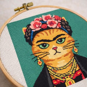 Könnte beinhalten: Kreuzstich-Porträt einer Katze, im Stil von Frida Kahlo, in einem hölzernen Stickrahmen. Die Katze hat orangefarbenes Fell, grüne Augen und trägt einen Blumenkranz, Ohrringe und eine Halskette. Der Hintergrund ist grün und weiß.