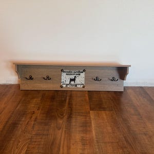 Könnte beinhalten: Ein hölzernes Wandregal mit vier schwarzen Haken und einem dekorativen Schild. Das Schild zeigt eine Hunde-Silhouette und den Text "RESERVE SUPREME MARKET DOG". Das Regal ist aus hellbraunem Holz gefertigt.