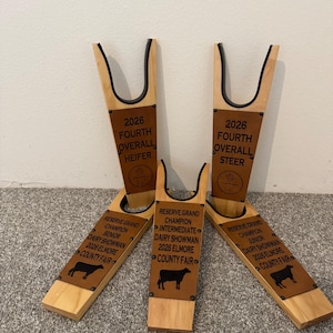 May include: Three wooden boot jacks with black rubber trim, each featuring engraved leather plates. The plates display text including 2026 Fourth Overall Heifer, 2026 Fourth Overall Steer, and Reserve Grand Champion Dairy Showman titles for the Elmore County Fair.