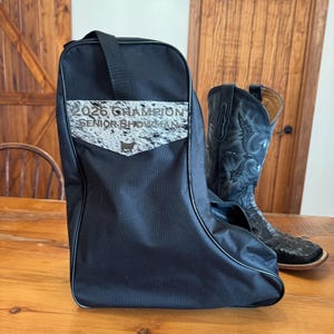 May include: Black boot bag with a shoulder strap and a black and white patterned pocket. The bag has the text "2026 Champion Senior Showman". A black cowboy boot with decorative stitching is next to the bag.